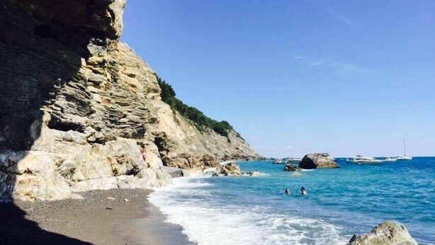 A coastal beach with rocky cliffs, clear blue water, and boats in the distance. - Le Pinette Bed and Breakfast (Ameglia)