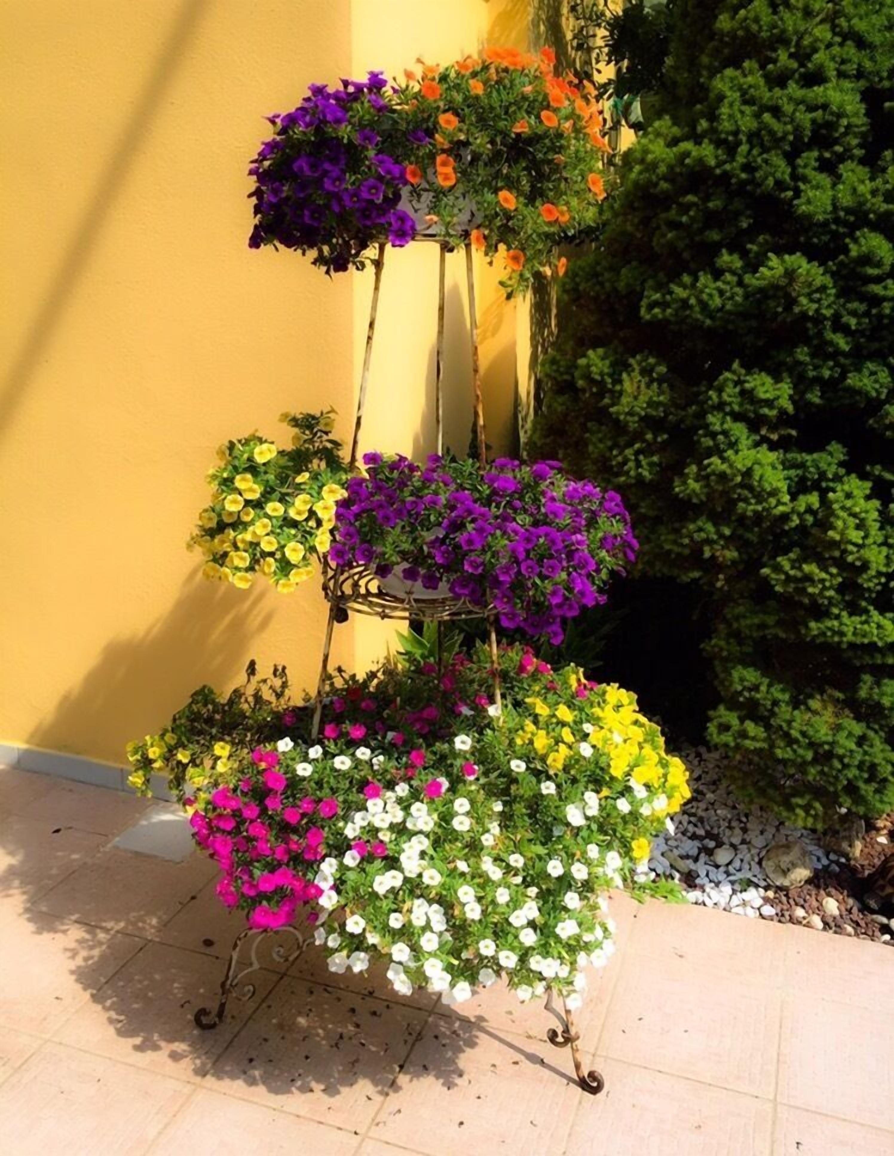 A tiered flower stand with various colorful flowers against a yellow wall and a green bush.