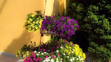 A tiered flower stand with various colorful flowers against a yellow wall and a green bush.