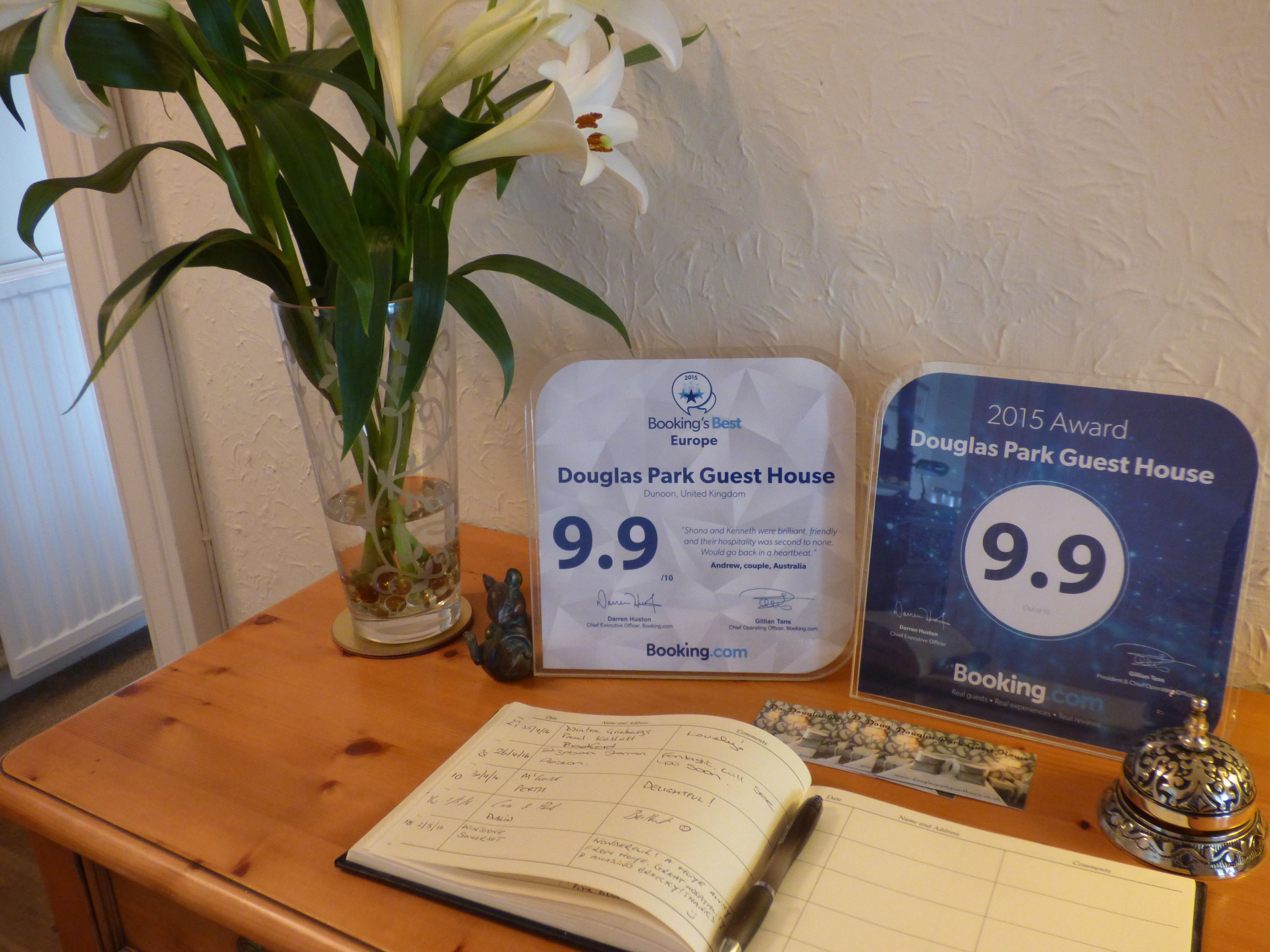 Una mesa de madera con un cuaderno, un bolígrafo, una campana decorativa, un florero de vidrio con una planta y dos placas de reconocimiento para Douglas Park Guest House.