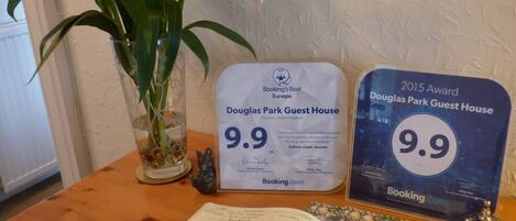 Una mesa de madera con un cuaderno, un bolígrafo, una campana decorativa, un florero de vidrio con una planta y dos placas de reconocimiento para Douglas Park Guest House.