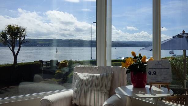 A room with a large window offering a view of a lake and distant hills. There is a striped armchair and a small table with a potted plant. A sign on the table indicates a price of 9.9. - Douglas Park Guest House (Dunoon)