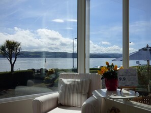 A room with a large window offering a view of a lake and distant hills. There is a striped armchair and a small table with a potted plant. A sign on the table indicates a price of 9.9. - Douglas Park Guest House (Dunoon)
