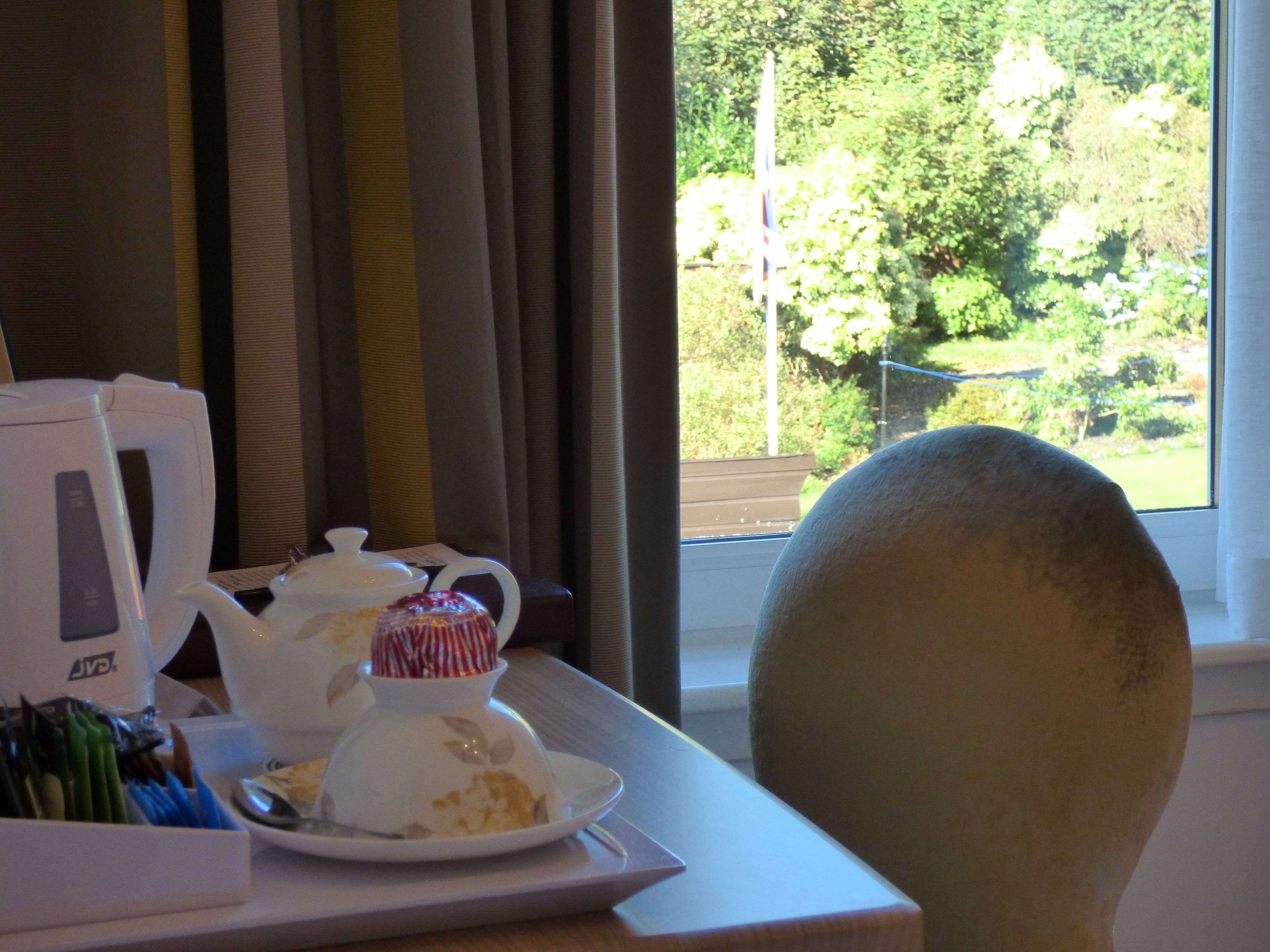 A table with a teapot, a cup, and a plate with food, a chair, and a window with a view of greenery.