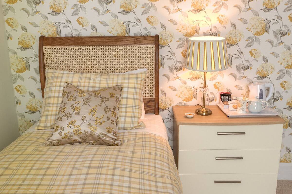 A bed with a checkered bedspread and floral pillows, a bedside lamp, a nightstand with a tray of books and a teapot, and a wall with floral wallpaper.