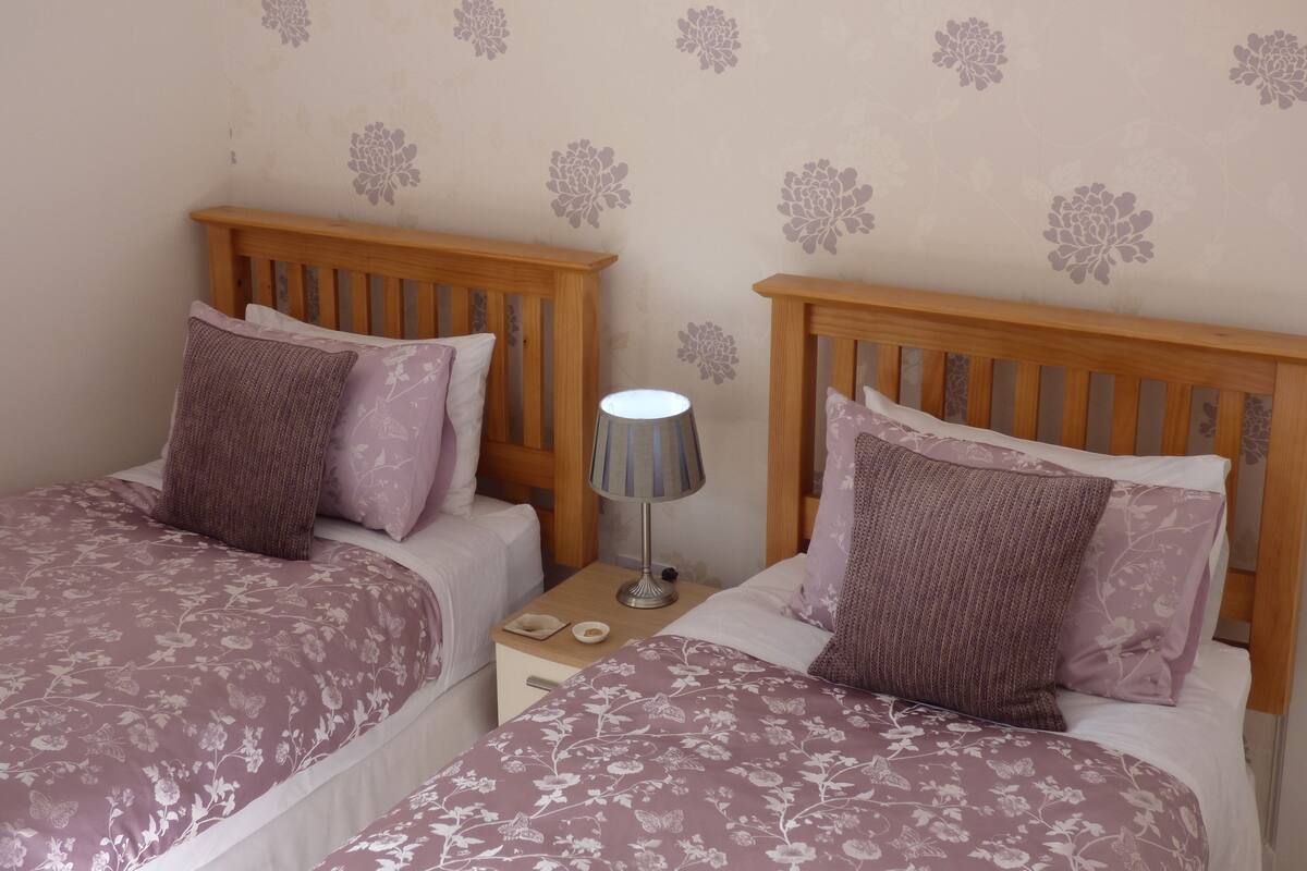 A room with two beds, each with floral-patterned bedding, a nightstand with a lamp, and a wall with floral decals.