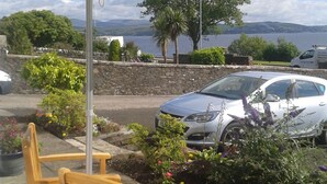 A wooden bench and table under a yellow umbrella, with a white car parked nearby and a scenic view of water and hills in the background. - Douglas Park Guest House (Dunoon)