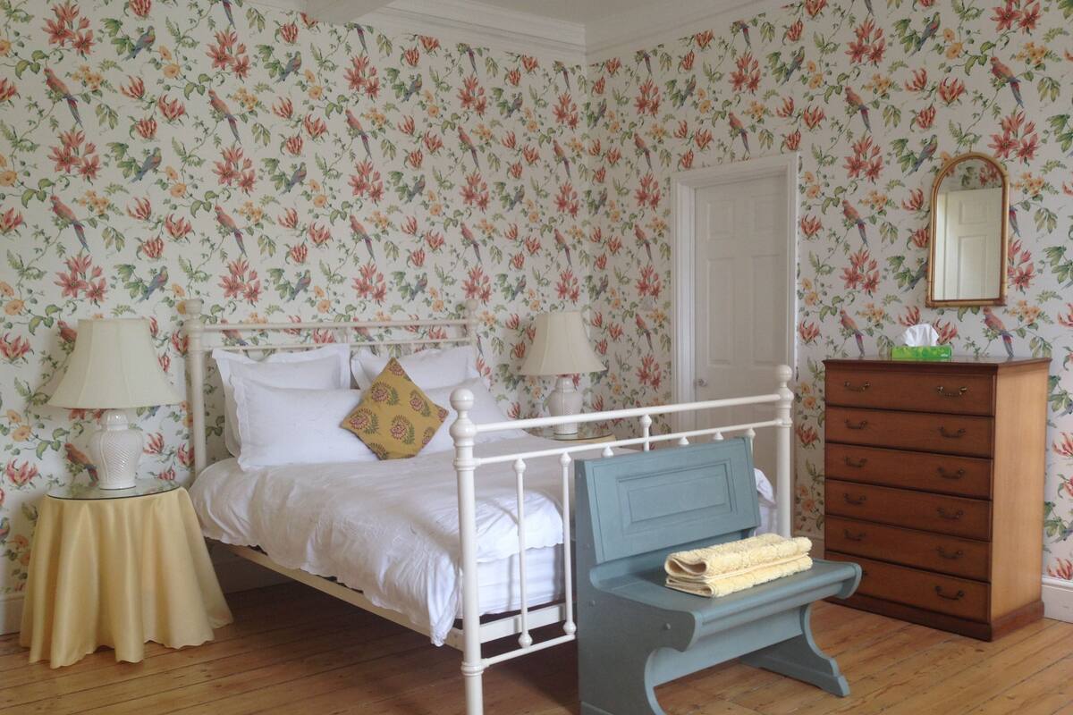 A bedroom with a white four-poster bed, a wooden dresser, a bench, and floral wallpaper.