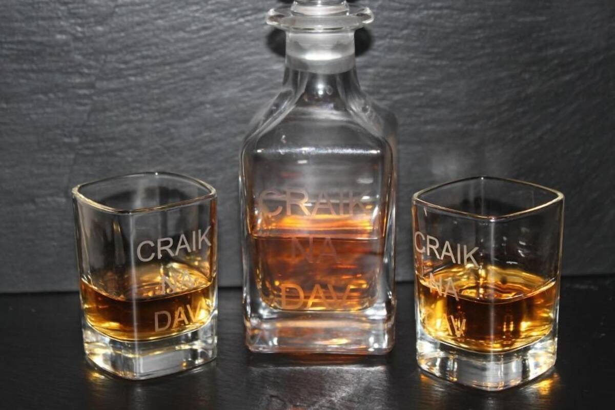 Ein Dekanter und zwei Schnapsgläser mit dem eingravierten Schriftzug "CRAIK DAV", auf einer dunklen Oberfläche platziert.