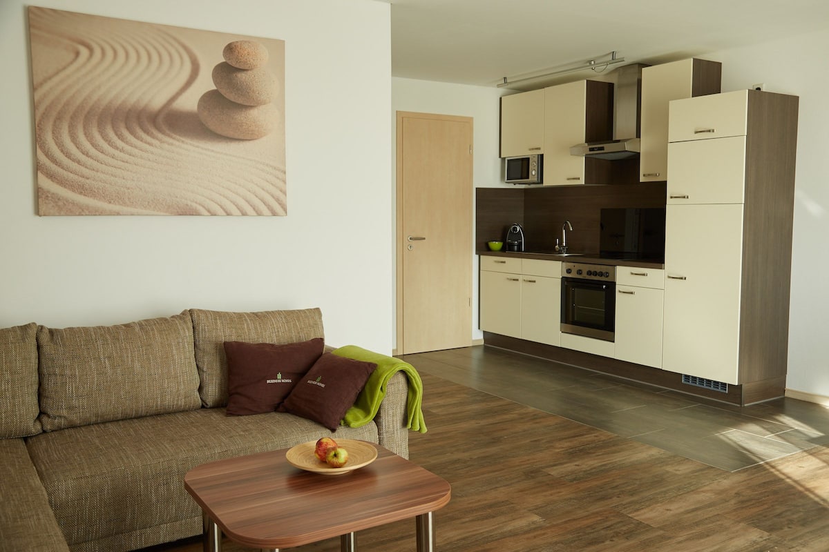 Een moderne woonkamer met een bank, een salontafel met fruit, en een keuken met kasten en apparatuur.