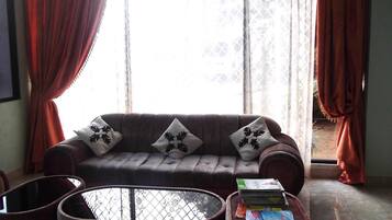 A living room with a brown sofa, patterned cushions, a glass coffee table, and a stack of magazines.
