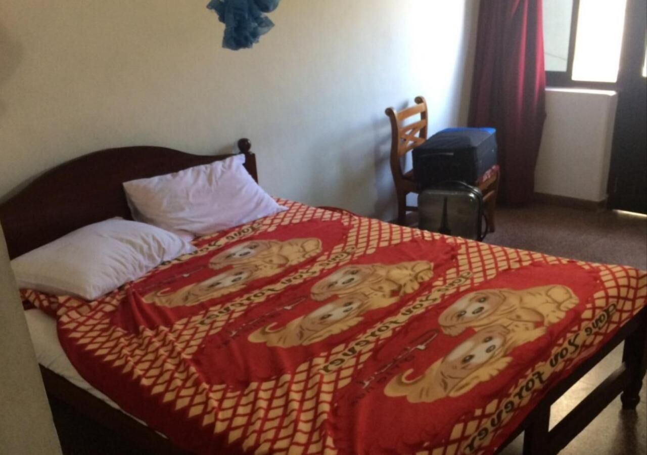 A bed with a patterned bedspread, a wooden headboard, and a chair in a room with a window and curtains.