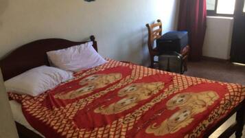 A bed with a patterned bedspread, a wooden headboard, and a chair in a room with a window and curtains.