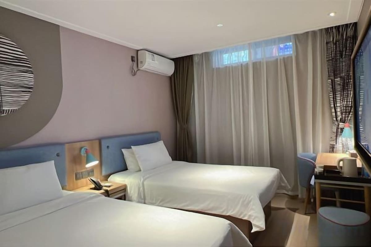 Ein Hotelzimmer mit zwei Betten, einem Schreibtisch, einem Stuhl, einer Lampe und einem mit Vorhängen versehenen Fenster.