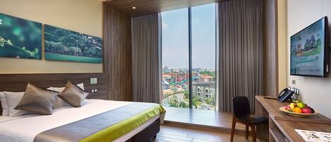 A hotel room with a large bed, a desk with a fruit bowl, a chair, and a view of a cityscape and water park through the window.