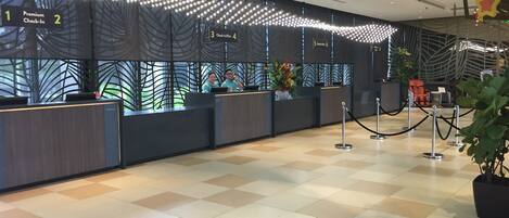 Zona de recepción de hotel con mostradores de registro de entrada, uno de salida y uno combinado. Hay luces decorativas y una planta en el centro.