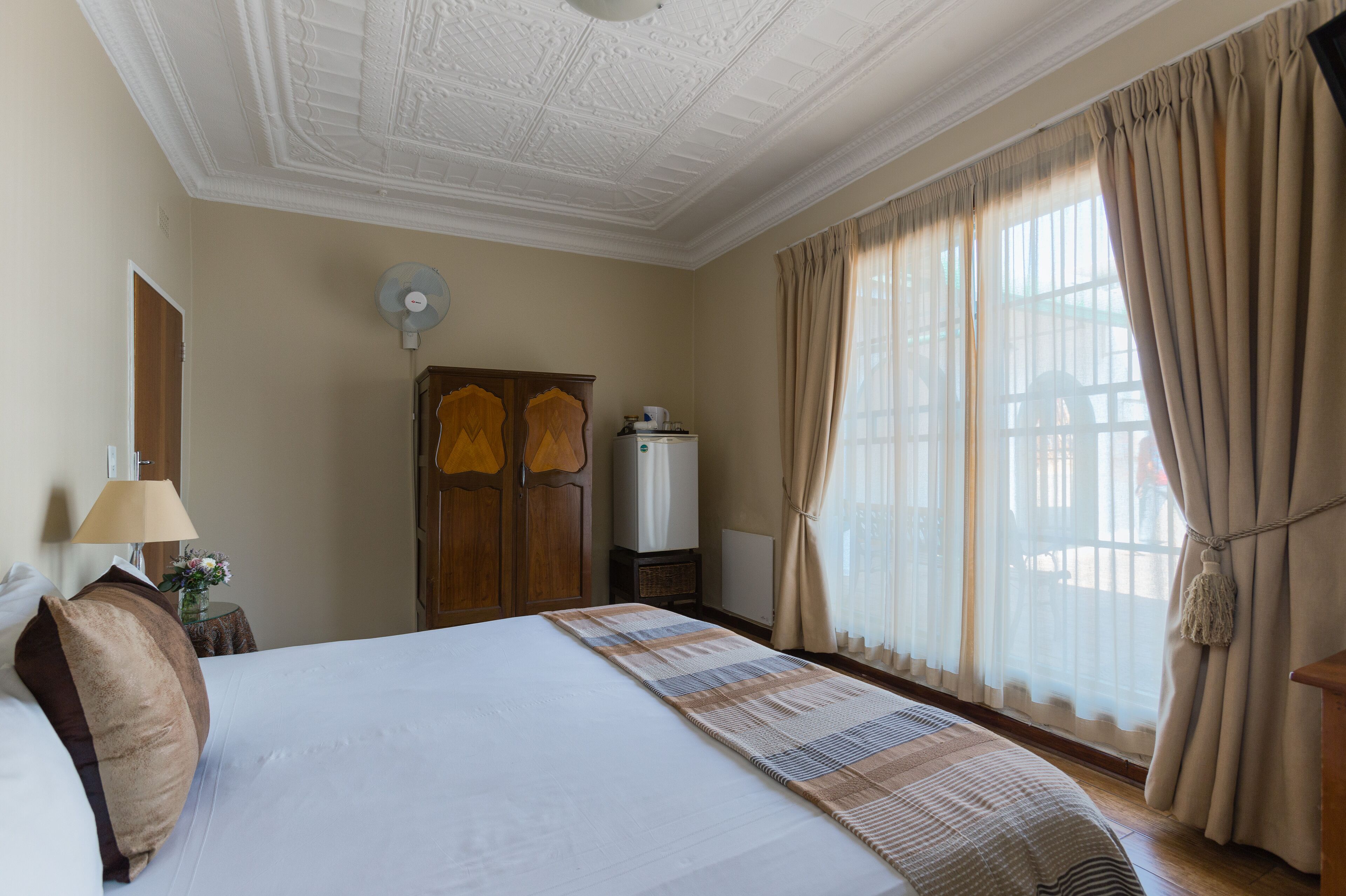Una habitación de hotel con una cama grande, un ventilador de techo, un radiador y una ventana con cortinas.
