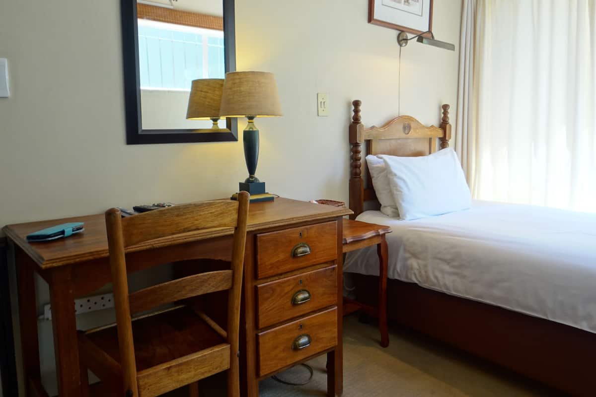A hotel room with a wooden desk, a lamp, a bed with white bedding, a framed picture on the wall, and a window with curtains.