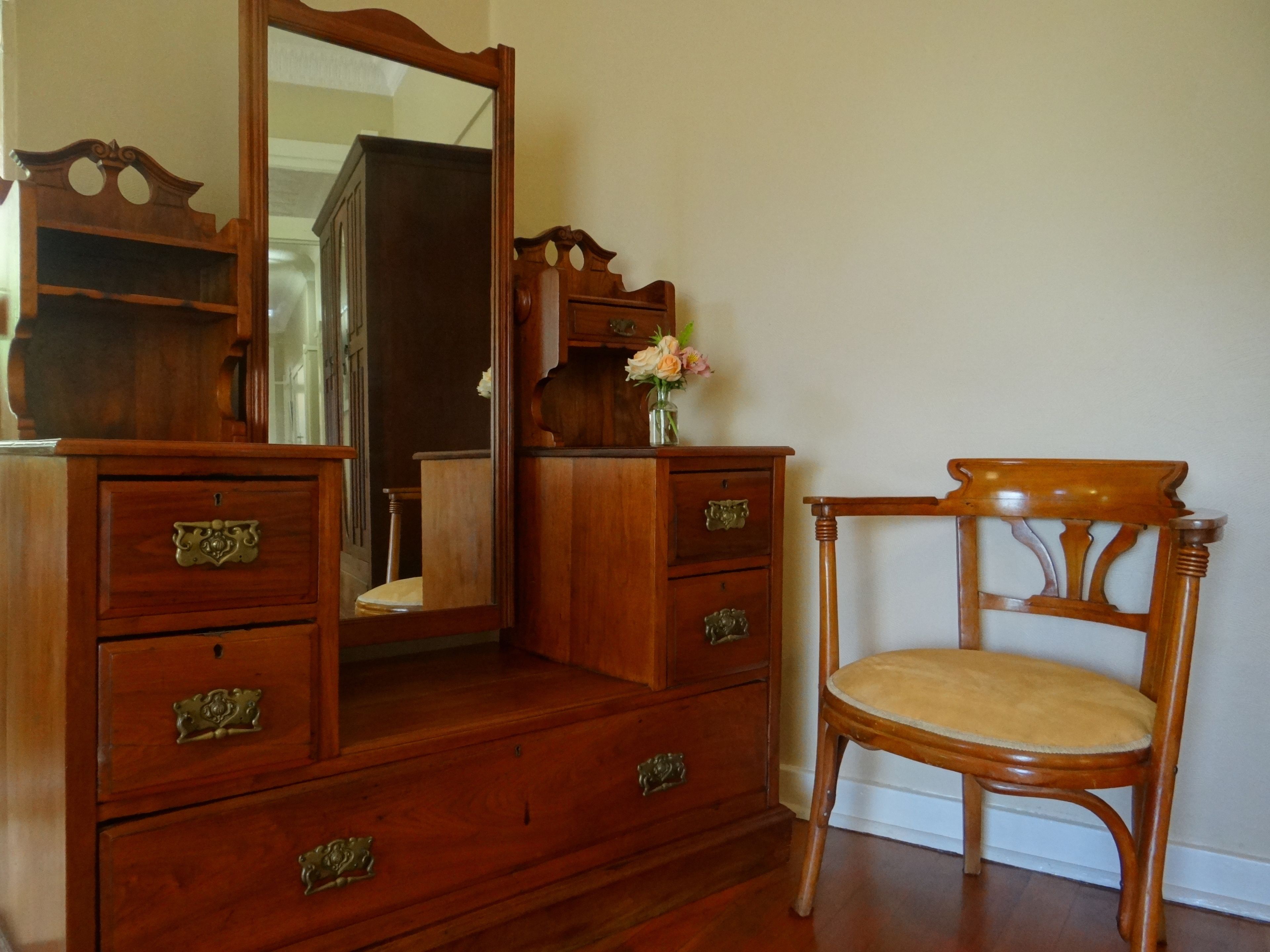 A wooden dresser with drawers and a mirror, a chair with a cushioned seat, and a vase with flowers on top of the dresser.