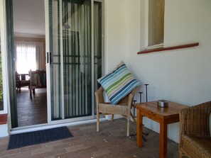 A room with a sliding glass door leading to a balcony, a wicker chair with a striped pillow, a wooden table, and a small metal container. - Agterplaas Guesthouse B&B (Randburg)
