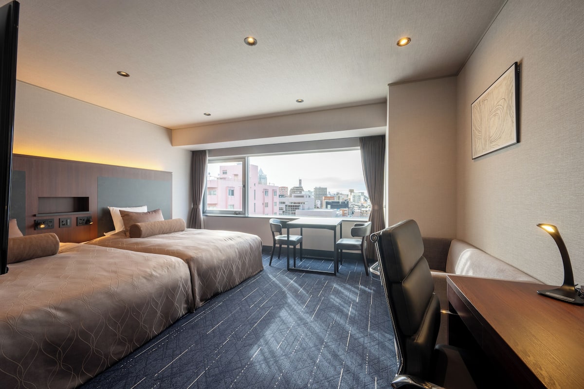 Ein Hotelzimmer mit zwei Betten, einem Schreibtisch und einem Stuhl. Ein Fenster bietet Blick auf die Stadt.