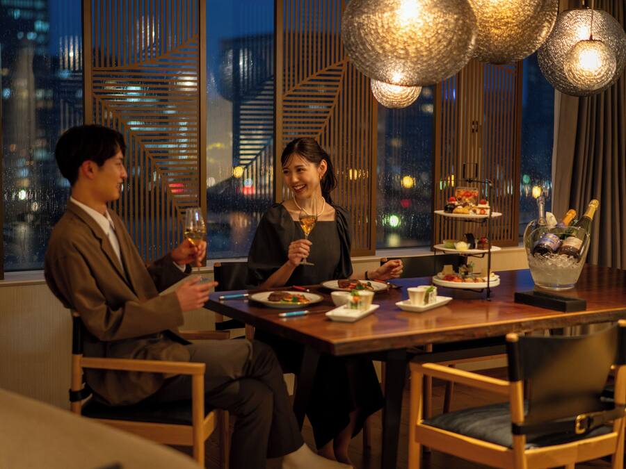 Una pareja cenando en una mesa con vistas a la ciudad, una botella de champán y una bandeja de postres.