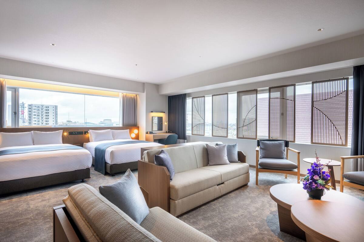 Ein modernes Hotelzimmer mit einem großen Bett, einer Sitzecke, einem Schreibtisch und Blick auf die Stadt durch große Fenster.