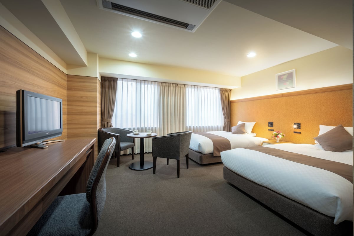 Ein Hotelzimmer mit Bett, Schreibtisch mit Fernseher, Sessel, kleinem Tisch und einem Fenster mit Vorhängen.