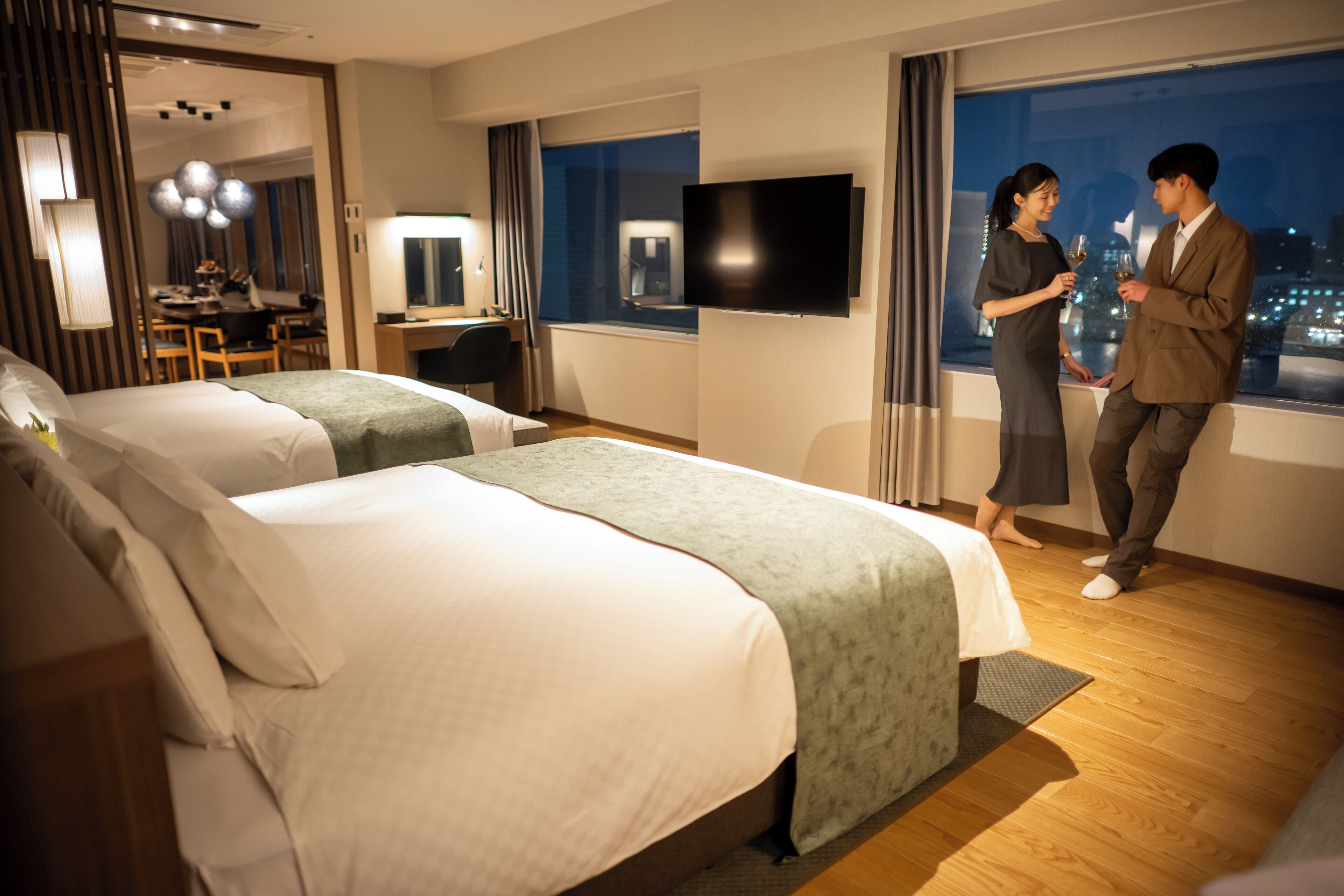 A hotel room with two beds, a desk, a TV, and a city view.