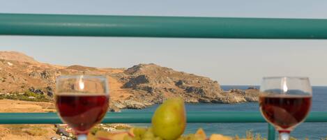 A table with a plate of fruit and two glasses of red wine, overlooking a coastal landscape with mountains and the sea.