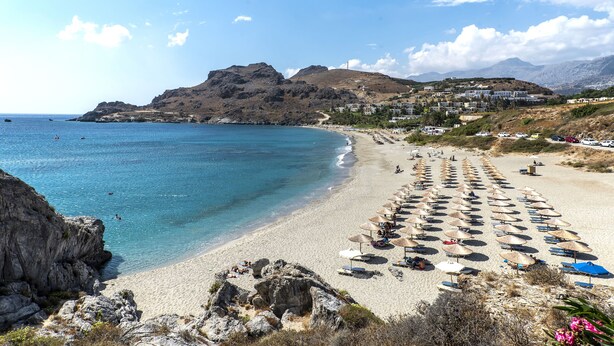 A beach with rows of sun umbrellas and beach chairs, a rocky coastline, and buildings in the background. - Sokol Resort (Agios Vasileios)