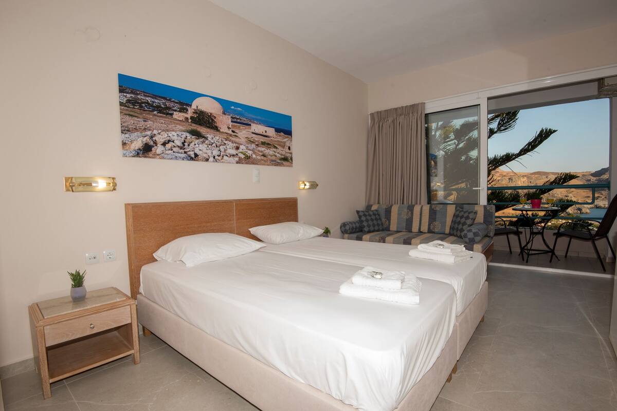 A hotel room with two beds, a small table with a plant, a painting of a landscape, and a balcony with chairs and a table.
