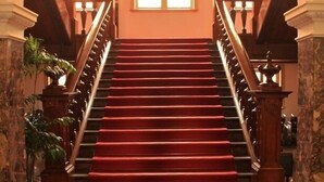 A grand staircase with red carpeted steps, flanked by ornate wooden railings and columns, leading to a bright room with large windows. - Yamagata Grand Hotel (Yamagata)