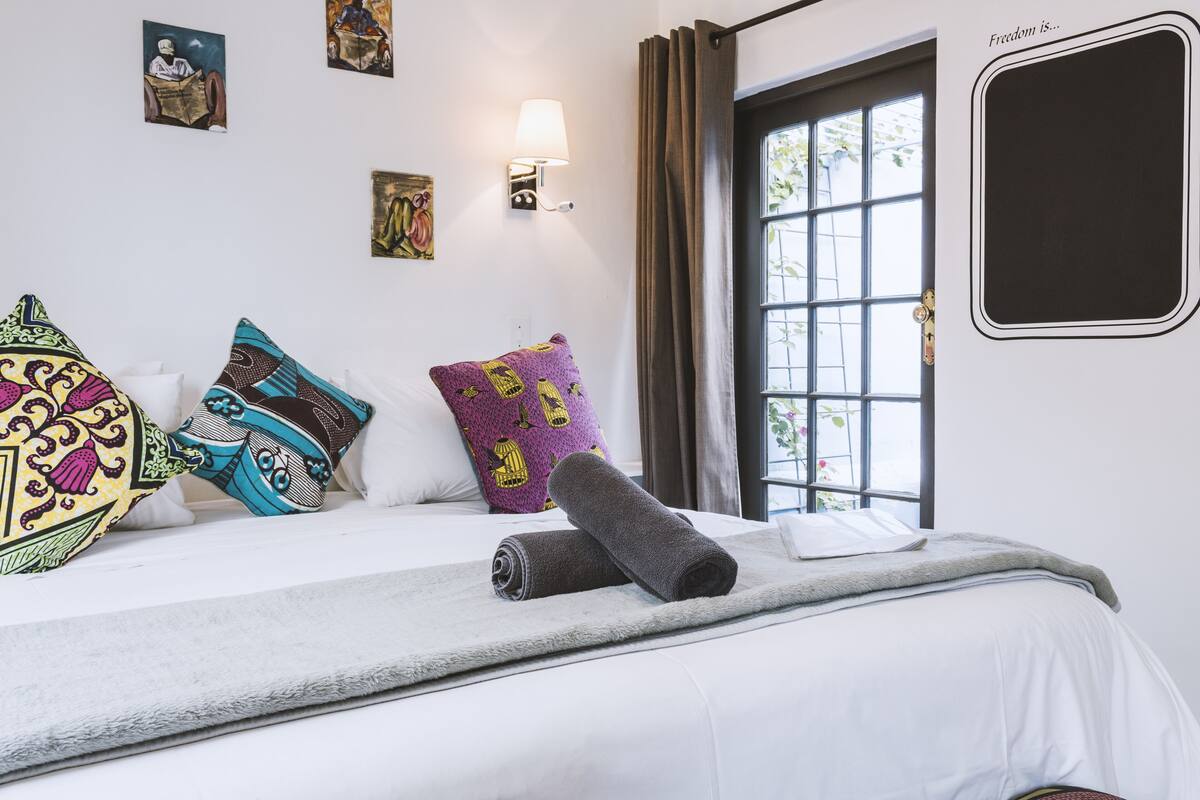 A neatly made bed with colorful pillows, a rolled towel, and a window with curtains.