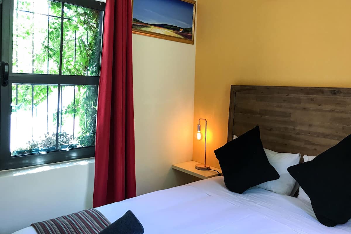 A hotel room with a bed, a nightstand, a lamp, a window with red curtains, and a view of greenery outside.