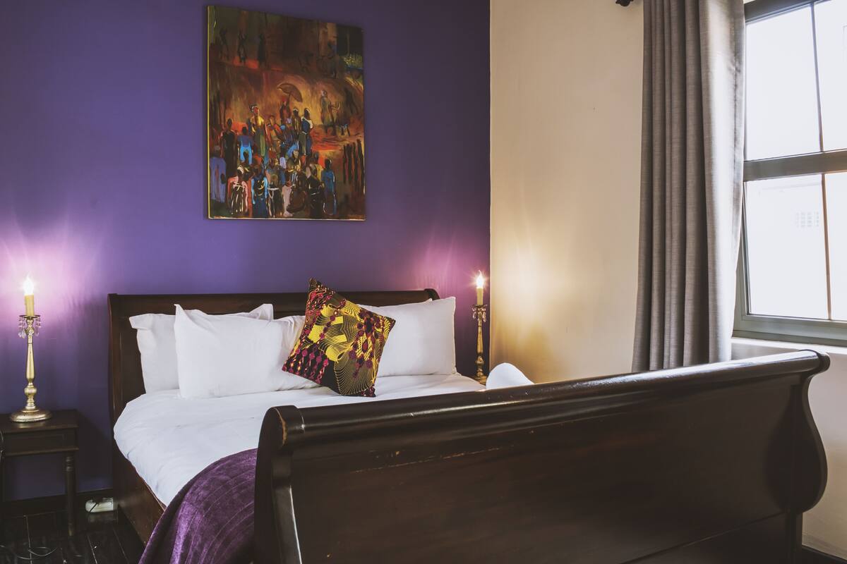 A bedroom with a large bed, purple walls, a painting above the bed, and a window with curtains.