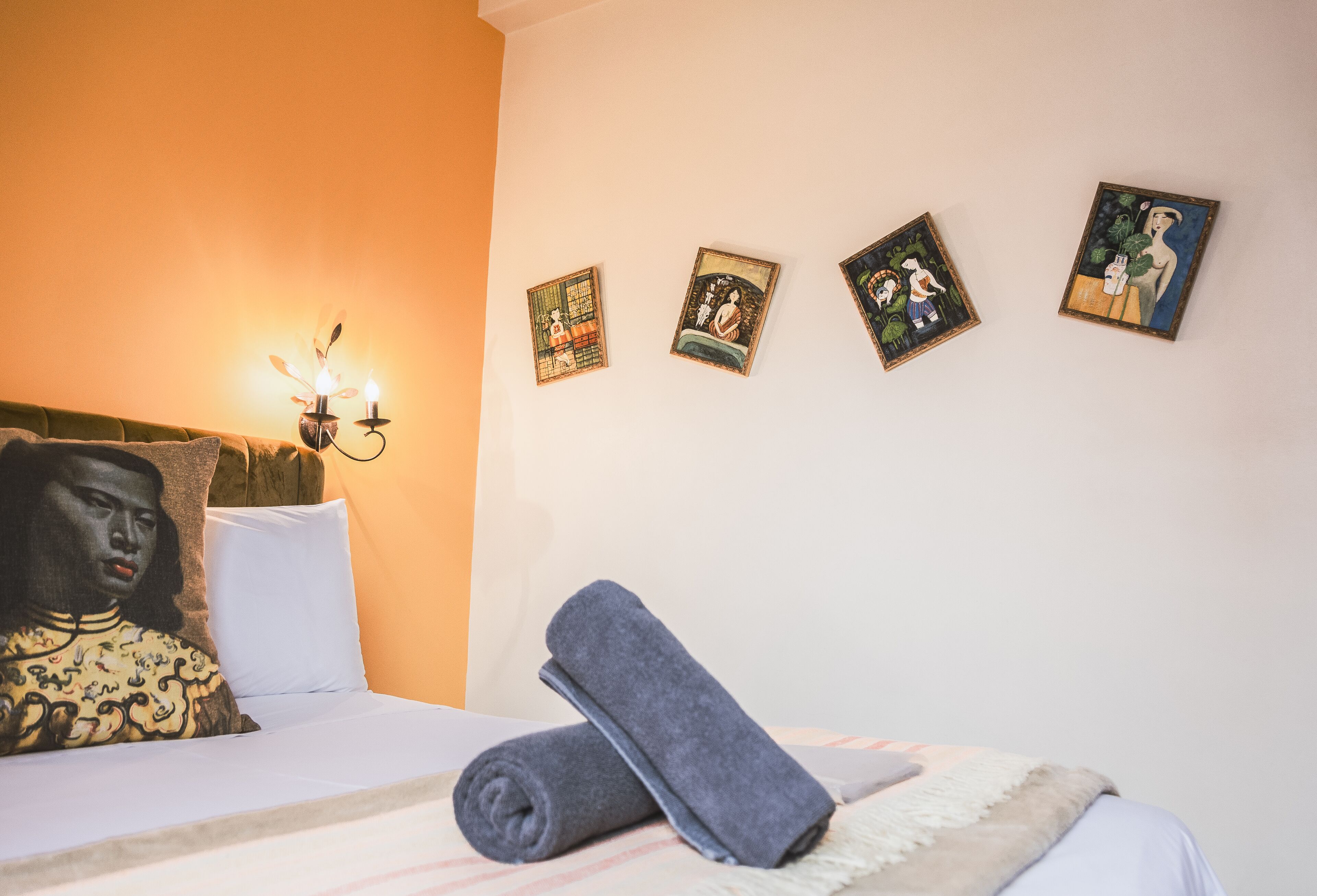 Uma cama bem arrumada com uma toalha enrolada, um travesseiro com desenho de rosto e quatro obras de arte emolduradas na parede.
