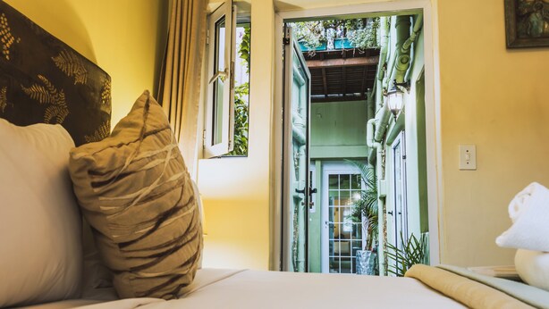 A bedroom with a bed, pillows, a window, and a view of a green outdoor area. - La Rose B&B (Cape Town)