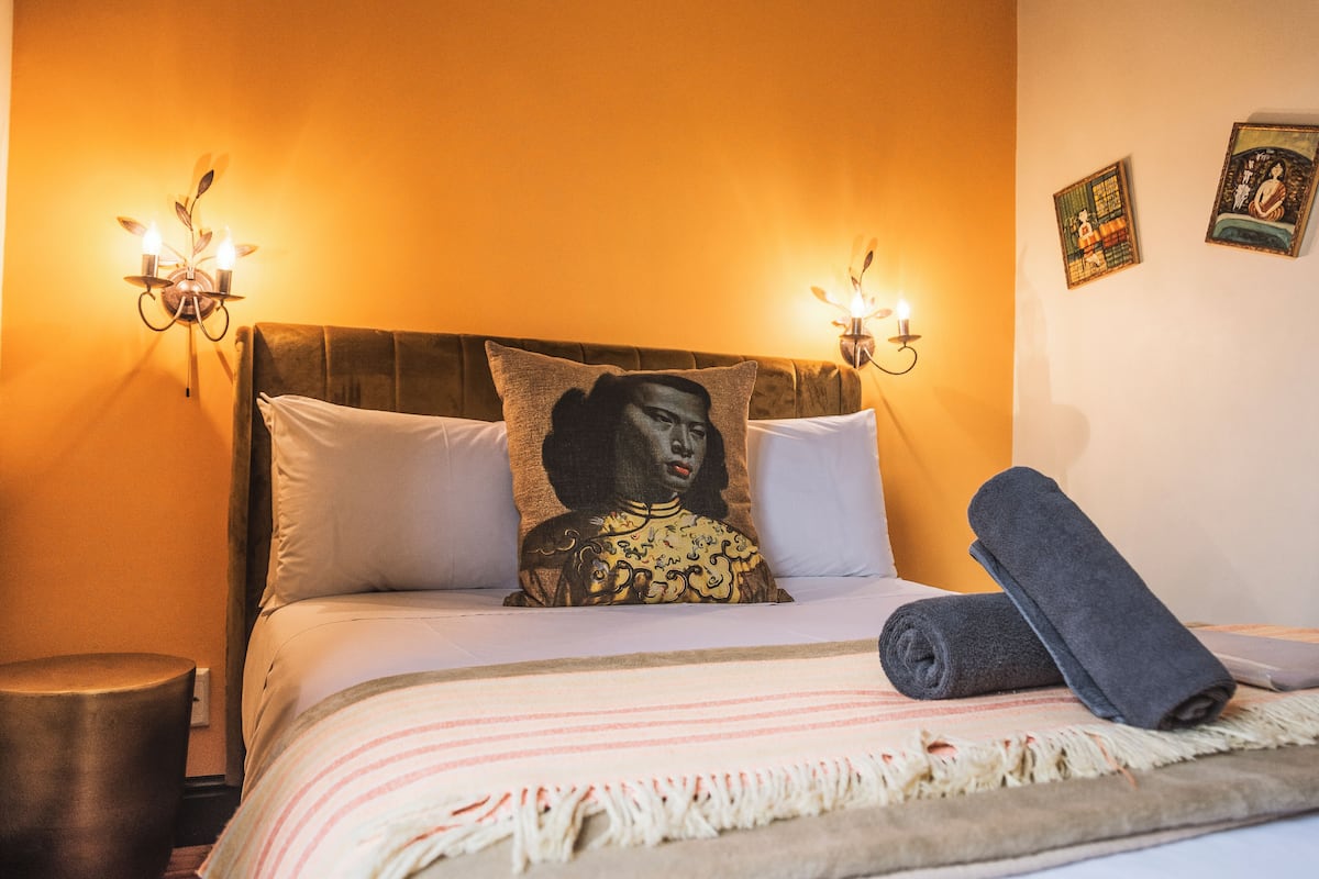 A neatly made bed with a pillow featuring a portrait, two wall-mounted lights, and a rolled towel on the bed.