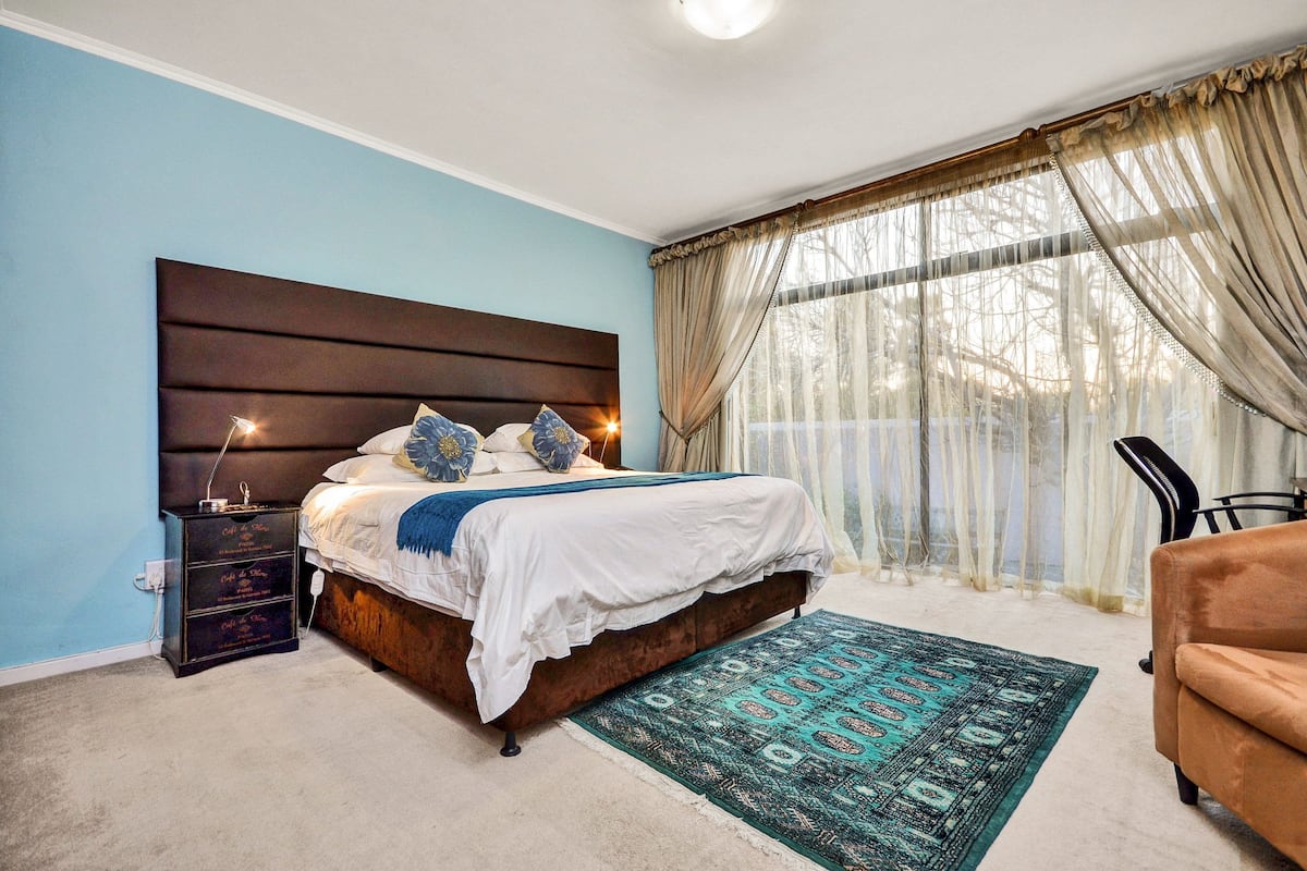 Quarto com cama grande, mesa de cabeceira, uma cadeira e vista para o exterior através de cortinas translúcidas.