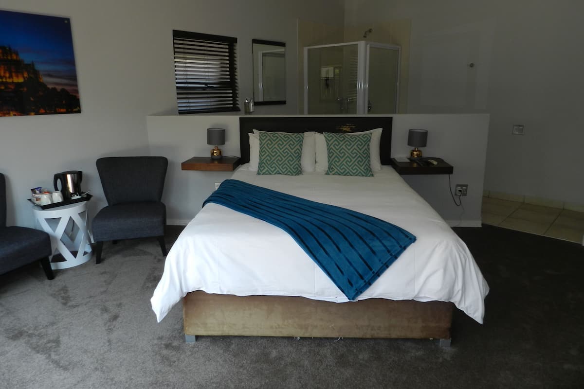 Quarto de hotel com uma cama grande, uma cadeira, uma mesa pequena com abajur e um espelho sobre a cama.