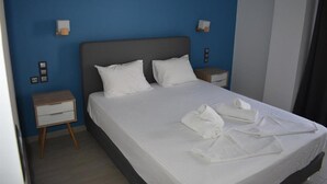 A neatly made bed with white linens and pillows, a bedside table with a lamp, and a blue wall. - Christina Apts and Studios (Corfu)