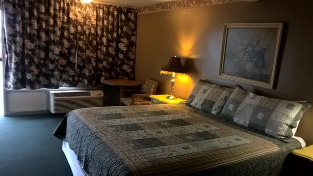 A hotel room with a large bed, a side table with a lamp, a chair, and a painting on the wall. - Dogwood Motel (Mountain View)