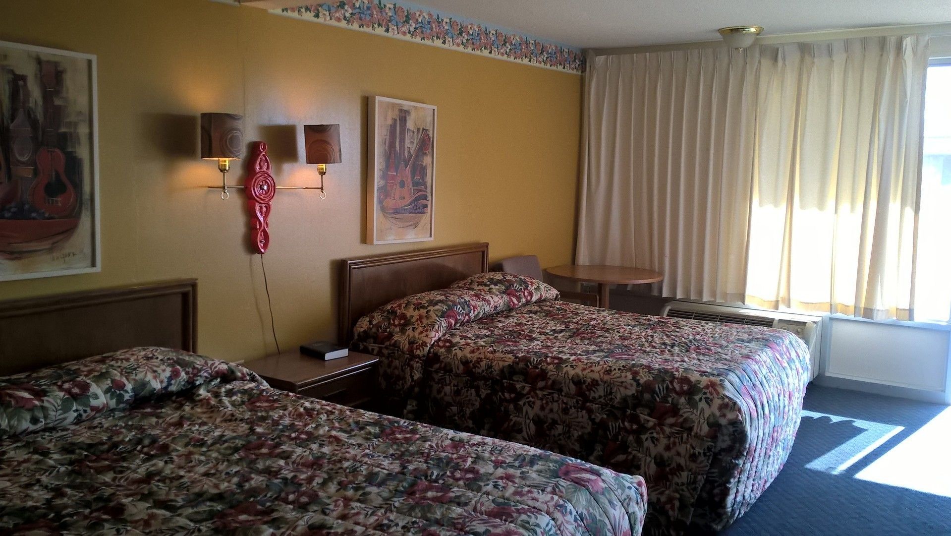 Une chambre d’hôtel avec deux lits, un bureau et une chaise. Les murs sont ornés de tableaux et il y a une fenêtre avec des rideaux.