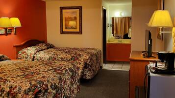 A hotel room with two beds, a desk with a lamp, a painting on the wall, and a bathroom visible through an open door.