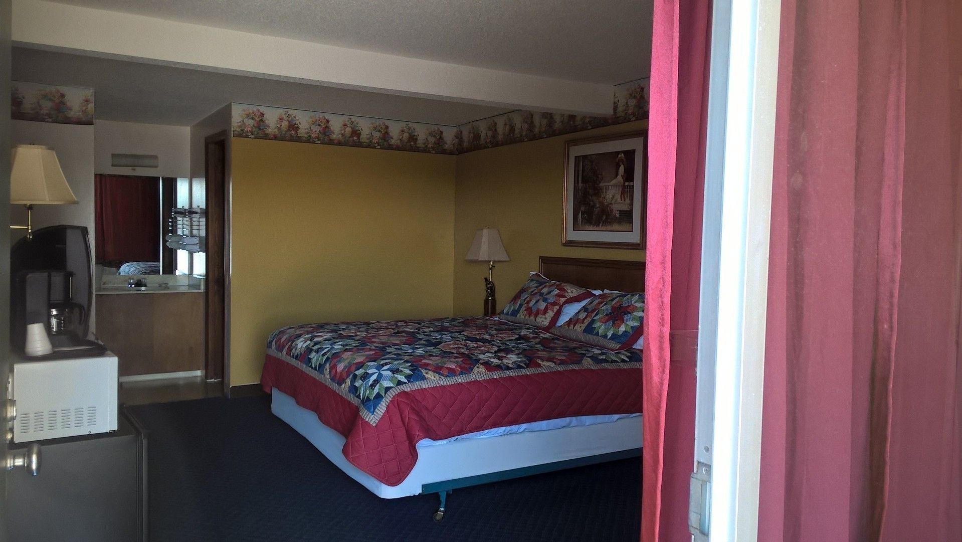 A hotel room with a bed, a desk with a lamp, a coffee maker, and a painting on the wall.