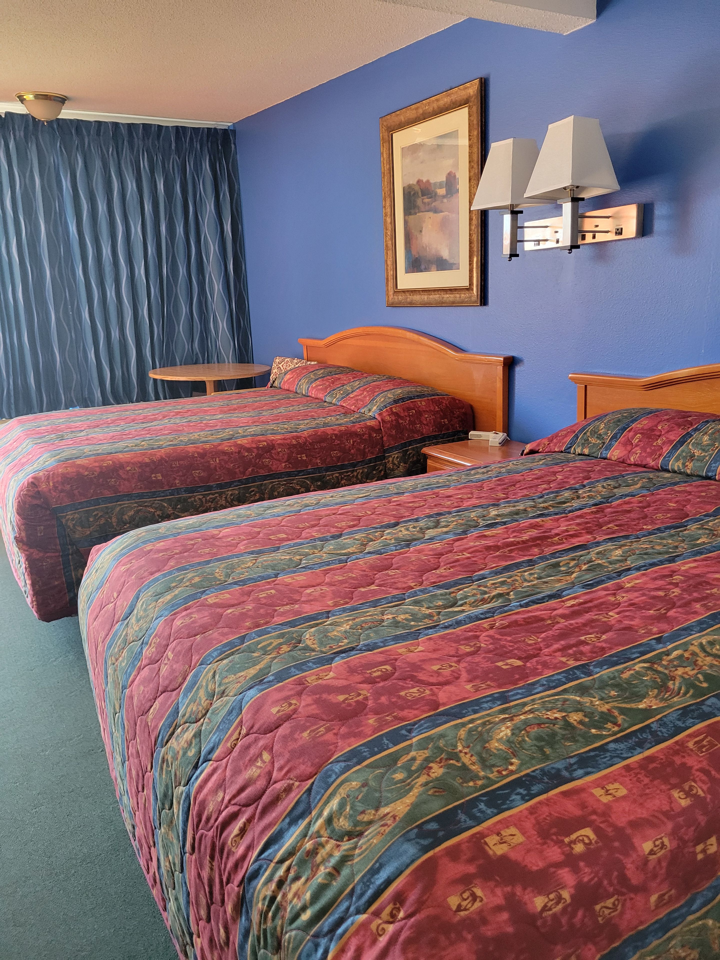 A hotel room with two beds, a round table, a lamp, and a painting on the wall.