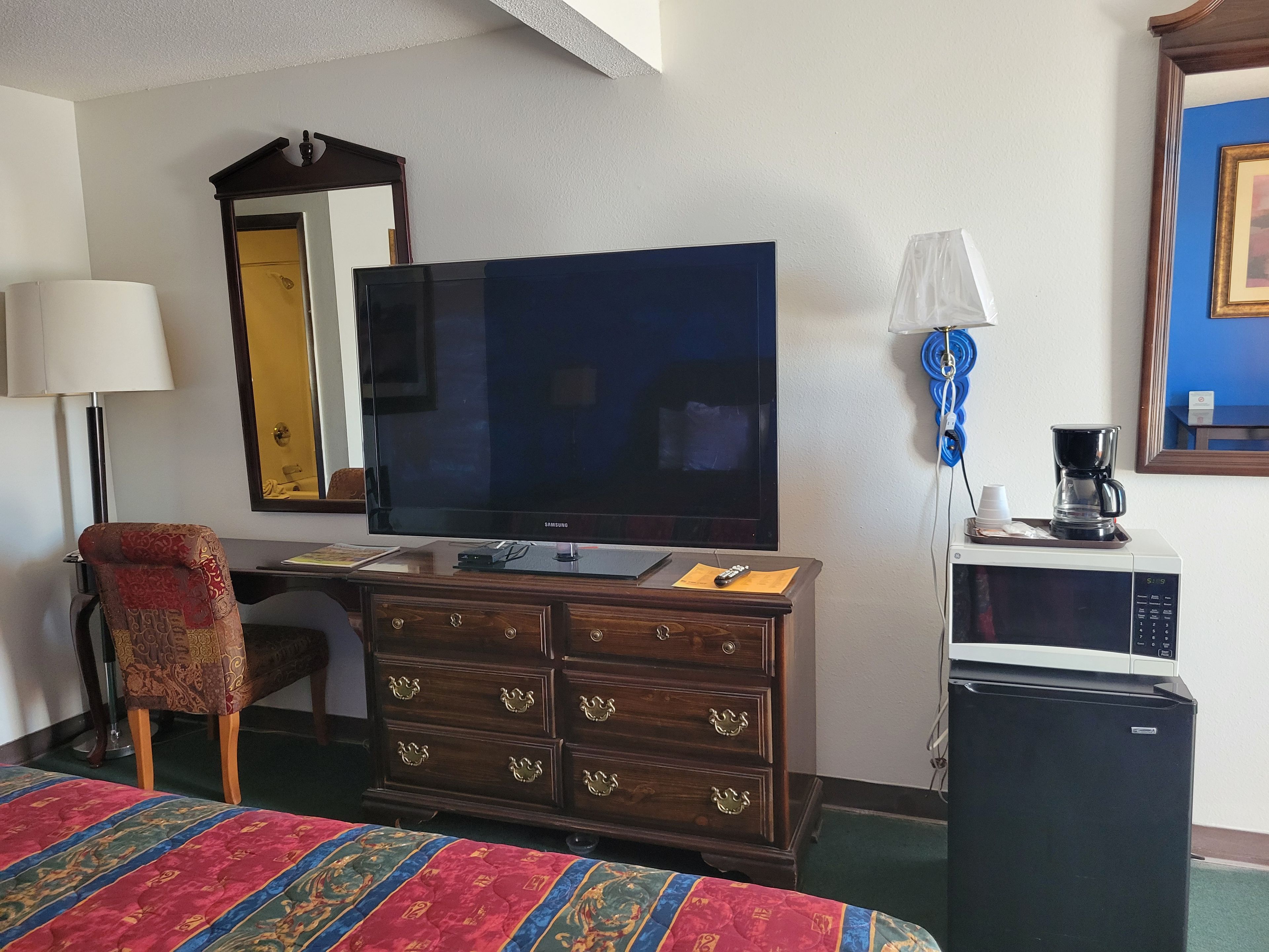 Ein Hotelzimmer mit Fernseher, Schreibtisch, Sessel, Kommode, einem Bett mit bunter Bettdecke, einer Kaffeemaschine und einem Kühlschrank.