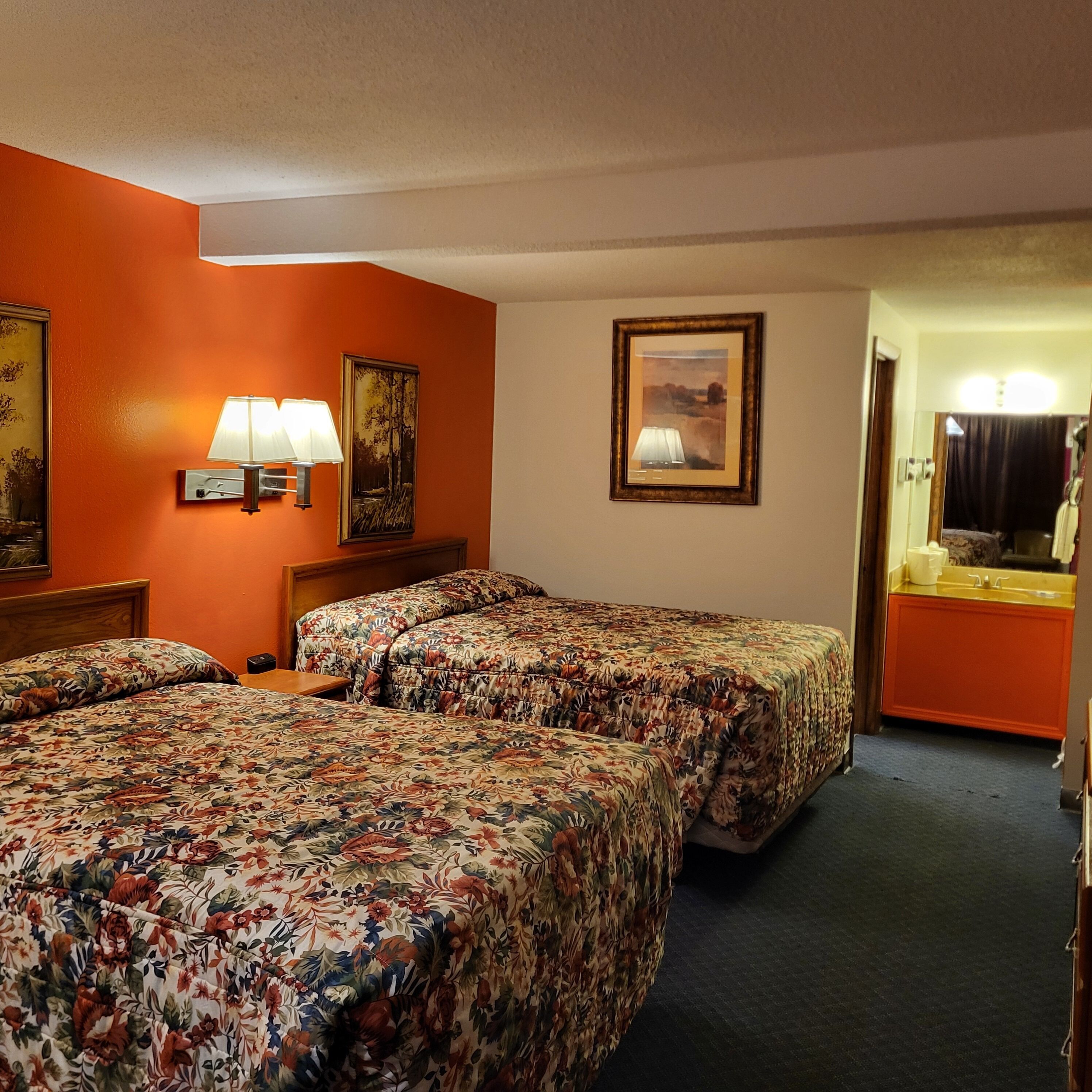 Une chambre d’hôtel avec deux lits, une table de chevet, une lampe, un tableau encadré et une salle de bain visible à travers une porte ouverte.