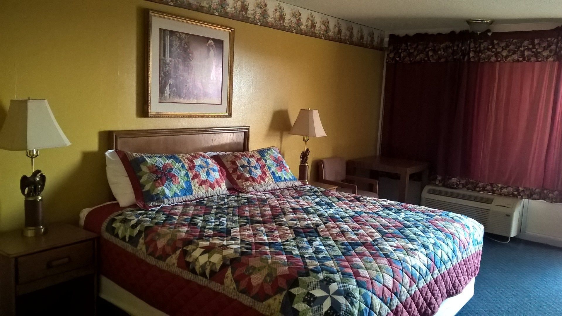 A hotel room with a large bed, two bedside lamps, a framed picture on the wall, a small table, and a window with curtains.